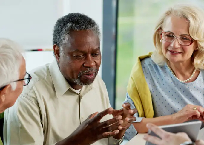 A group of two older men and an older woman sit together having a conversation. The man in the middle uses his hands to talk while the three look at something being displayed on a screen in front of them.