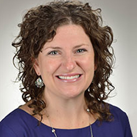 A woman with brown curly shoulder-length hair smiling in a blue shirt with jewelry on. In front of a plain grey background.