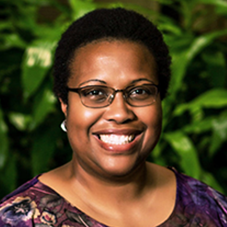 A woman with short dark hair and dark framed glasses in a purple patterned shirt smiling. She is in front of a background with greenery.