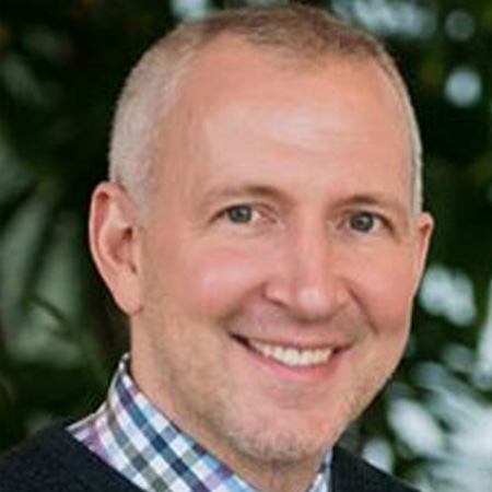 A man with short gray hair smiling wearing a plaid collard shirt. He is in front of a background with greenery.