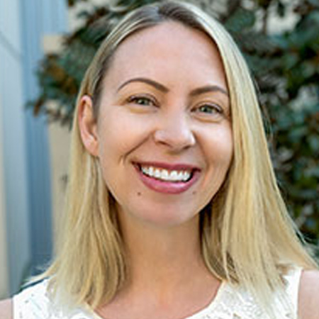 A woman with shoulder length blonde hair in a white shirt smiling. She is in front of tree.
