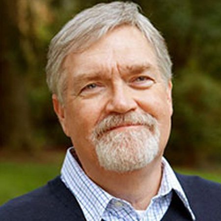 A man with short gray hair and a gray beard smiling wearing a blue plaid collard shirt. In front of a greenery background.